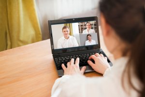 A woman sits at a wooden desk with a laptop, participating in a remote video conference. The screen shows three people in separate video frames, engaging in conversation. The background includes curtains and a window with natural light.