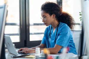 A healthcare professional wearing blue scrubs and glasses sits at a desk, intently focused on typing on a laptop. In the foreground, various medical supplies and documents are neatly arranged. The background features large windows with natural light streaming through, highlighting the serene atmosphere of the healthcare office.