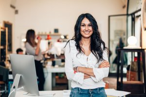 A confident leader with long dark hair, wearing a white blouse and jeans, smiles with her arms crossed while standing in an office. A computer monitor is on a desk in front of her, and there are blurred colleagues in the background.
