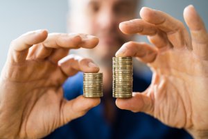 A person holding two stacks of coins side by side. The stack on the left is shorter than the stack on the right. Gender is not apparent, as the focus is solely on the hands and coins, with a blurred background enhancing this effect.