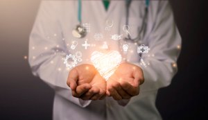 A person in a white lab coat and stethoscope holds glowing, doodle-like medical icons in cupped hands. The icons form a heart shape, symbolizing healthcare and care. The background is dark, highlighting the illuminated icons and hands.