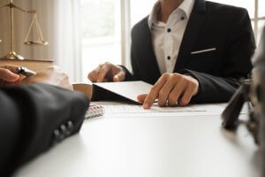 Two business professionals in formal attire are engaged in a meeting at a desk. One person gestures toward a document with a pen, while the other follows along. Scales of justice and books are seen in the background, indicating a legal or government office setting.