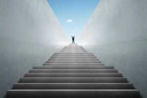 A person stands with arms raised in triumph at the top of a long flight of wide, concrete stairs. The stairs, symbolizing development, are flanked by high, smooth concrete walls that converge towards a bright blue sky with a single white cloud.