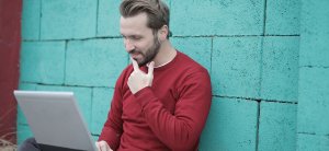 A man with a beard and short hair, wearing a red sweater, is sitting against a turquoise brick wall, working on his laptop. Resting his chin on his hand, he appears deep in thought as he pores over "The Ultimate Guide to Finding Jobs in DC Government" displayed on the screen.