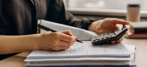 A person in a dark shirt uses a pen to write on documents on a desk while holding a calculator. A coffee cup and other office supplies are visible in the background near a window, with "The Ultimate Guide to Finding Jobs in DC Government" booklet resting beside them.