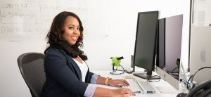 A woman with long, wavy hair sits at a desk in an office, smiling at the camera. She is working on a computer with dual monitors, one of which displays "The Ultimate Guide to Finding Jobs in DC Government." Behind her, a whiteboard features some written notes and diagrams.