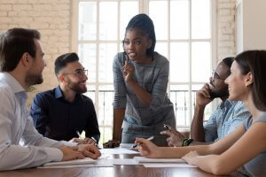 A diverse group of five colleagues in a meeting room engage in a discussion at a table. A woman, clearly the leader, stands speaking with confidence as the others sit and listen attentively. The room has a large window bringing in natural light. Notepads and documents are on the table.