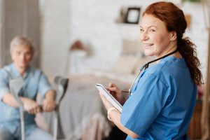 A smiling healthcare professional in blue scrubs and a stethoscope holds a clipboard, consulting with a patient seated in the background using crutches in what appears to be a comfortable living space.