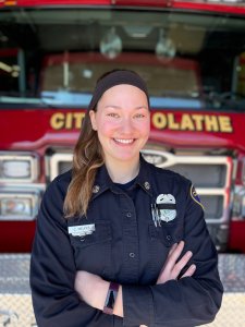 A firefighter in uniform stands confidently with her arms crossed in front of a red fire truck. She is wearing a headband and a name tag on her chest. The words "City of Olathe" are visible on the fire truck behind her.