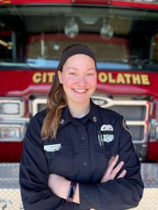 A smiling firefighter in uniform stands with arms crossed in front of a red fire truck. The truck has "CITY OF OLATHE" written on it.