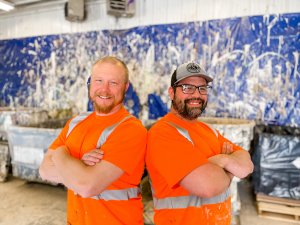 Two men stand back-to-back with their arms crossed, smiling at the camera. They are wearing bright orange shirts with reflective stripes and glasses. The background features a wall with abstract paint splatters in various colors.