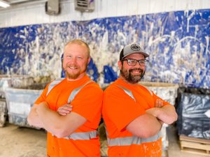 Two men wearing bright orange safety shirts and glasses stand back-to-back with arms crossed, smiling, in a workshop with a splattered paint backdrop.