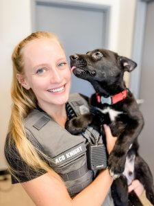 A woman in uniform, with the name "ACO Knight" on her vest, smiles while holding a black puppy with a red collar. The puppy is licking her face. They are indoors with a gray door and wall in the background.