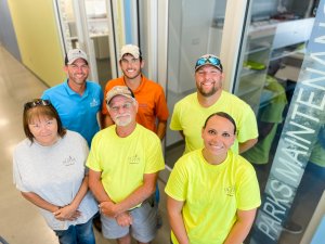 A group of six people wearing casual work clothes stands and smiles in a hallway. Three men and one woman are in bright yellow shirts, while another man and woman wear light gray and blue shirts, respectively. A "Parks Maintenance" sign is visible on the glass door behind them.