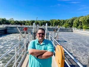 A man in a light blue polo shirt stands with arms crossed on a metal walkway overlooking a water treatment facility. Behind him are large circular tanks of aerated water. The background features green trees under a clear blue sky.