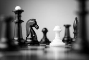 Black and white image of a chessboard with a white pawn in the foreground, symbolizing progress amidst black chess pieces, including a knight, rook, and bishop. The background is blurred, emphasizing the white pawn as the focal point.
