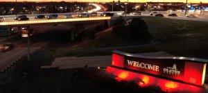 A nighttime scene showing a well-lit "Welcome to Olathe" sign in the foreground, illuminated in red. In the background, cars travel along a highway and a bridge, with city lights and buildings visible under a darkening sky.