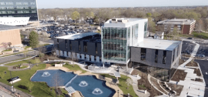 Aerial view of a modern building with large glass windows, surrounded by parking lots and green spaces. In front, there is a landscaped area with a pond featuring three fountains. Trees and other buildings can be seen in the background.