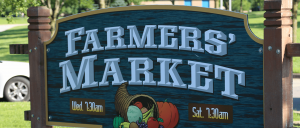 A wooden sign for a Farmers’ Market displays market hours—Wednesdays and Saturdays at 7:30am—and features a cornucopia filled with fruits and vegetables. Trees, grass, and a parked car are visible in the background.