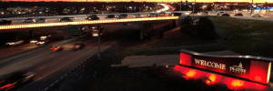 Nighttime highway scene featuring a lighted overpass with cars moving in both directions. A lit welcome sign for Olathe is seen on the right, with a glow of red lights beneath it. The distant horizon shows a glowing skyline as cars stream along the roads.