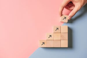 A hand places a wooden block with an upward arrow on top of a staircase structure made of similar blocks. The background is split diagonally into two colors: pink on the top left and blue on the bottom right, symbolizing governmental progress or growth.