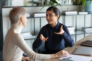 Two leaders are engaged in a conversation at a desk in a modern office setting. One woman has short, blonde hair and wears a white turtleneck. The other has dark hair tied back and wears a navy blue, polka dot blouse. A shelf with books and plants is in the background.