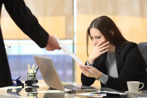 A woman sits at her desk with a laptop, looking worried and covering her mouth with one hand. The other hand holds a document, possibly a firing notice, handed to her by a person standing off-frame. Office supplies and paperwork are scattered on her desk.