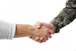 A handshake between two people is shown in this image. One person is wearing a military camouflage uniform, symbolizing duty and service, while the other is in a casual grey long-sleeve shirt. The background is white and unobtrusive.