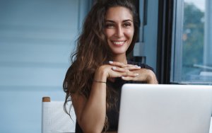 A smiling person with long, wavy hair sits at a desk with their hands clasped in front of a laptop, possibly working on government-related tasks. They are wearing a dark top, and a window with a view of greenery is visible in the background.