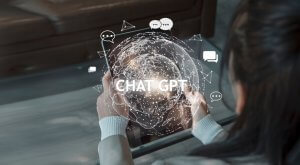A person holding a tablet displays a glowing digital globe with various icons and speech bubbles surrounding it. The text "ChatGPT" is prominently featured over the globe, indicating the use of AI chat technology. The background includes a desk and some furniture.