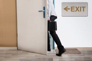 A person carrying a black briefcase is seen mid-step as they exit through a door, perhaps after a firing. The person's upper body is not visible. A sign with an arrow pointing left and labeled "EXIT" is mounted on the wall next to the door. The floor is covered with a light-colored mat.