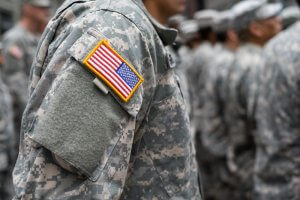 A close-up of a soldier wearing a camouflage military uniform with a U.S. flag patch on the sleeve. More soldiers in similar military uniforms are seen standing in formation in the blurred background.