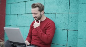 A man with light brown hair and a beard wearing a red sweater sits against a textured teal wall while using a laptop. With one hand thoughtfully touching his chin, he seems focused on searching for government jobs in Tarrant County.