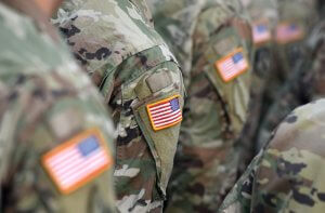 Several military soldiers wearing camouflage uniforms, each adorned with a U.S. flag patch on the arm, stand in formation. The focus is on the flag patches, emphasizing a sense of unity and patriotism.
