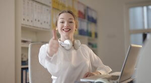 A smiling person wearing white headphones around their neck gives a thumbs-up gesture while sitting at a desk with a laptop. The background features shelves with colorful binders and folders, possibly hinting at their organization skills—perfect for government jobs in Riverside County.