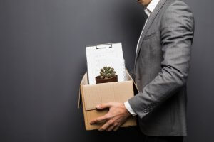 A person in a gray suit holds a cardboard box containing a potted cactus, a clipboard with papers, and a notebook, hinting at packing up their belongings due to being fired. The plain gray background adds to the somber mood of leaving their job.