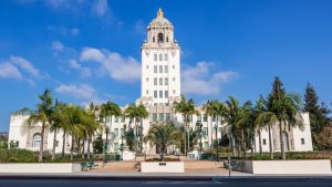 A large, ornate white building dedicated to public service stands with a tall central tower topped by a golden dome. The facade features columns and decorative details. Palm trees, flowers, and benches are in the foreground, set against a clear blue sky backdrop.