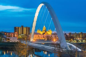 A modern arch bridge illuminated at evening spans over a calm river, leading towards a distant cityscape featuring a prominent illuminated historic building. The sky is a deep blue, enhancing the reflections and lights of the city, embodying the essence of public service in its infrastructure.