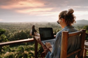 A person sits on a wooden chair on a balcony, working on a laptop with legs stretched out on the railing. They overlook a lush green landscape, under a cloudy sky with pink and orange hues at sunset. The person has their hair up and wears casual clothes and sunglasses, perhaps drafting an important government report.