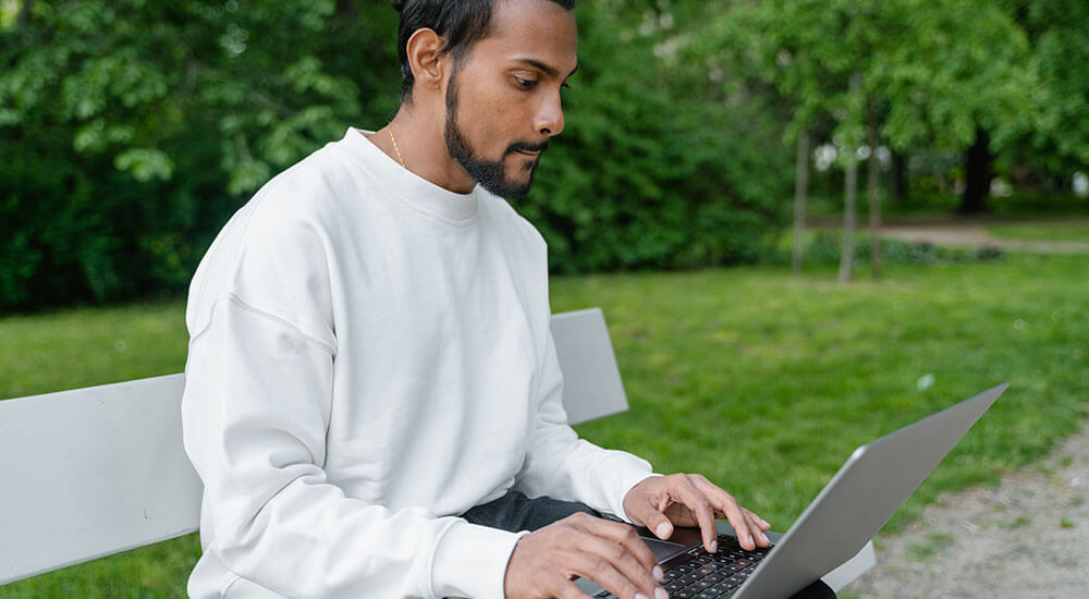 A man pressing a laptop while sitting in the park - New York State Park - Careers In Government