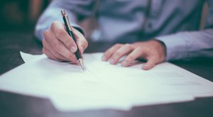 A person in a light blue shirt is diligently writing on white papers with a pen at their desk, possibly applying for government jobs in Collin County.