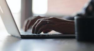 Close-up of hands typing on a laptop keyboard, possibly searching for government jobs in Collin County. One hand is wearing a ring. The background is softly lit, suggesting an indoor setting.