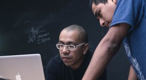 Two individuals are focused on a laptop. One person is wearing glasses and has a concentrated expression, while the other leans over, also looking at the screen—perhaps searching for government jobs in Collin County. A blackboard with faint writing is visible in the background.