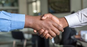 Two people are shaking hands in a well-lit, modern office setting, possibly discussing Government Jobs in Collin County. One person is wearing a light blue shirt, and the other is in a white shirt. In the background, there are blurred figures and furniture, indicating a professional environment.