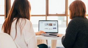 Two people sit at a white table, viewing a laptop screen displaying an open website about government jobs in Collin County. Both individuals have long hair and are facing away from the camera. Sunlight streams in from large windows in the background, illuminating the scene.