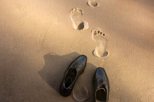 A pair of black dress shoes is placed on sandy ground with two sets of human footprints extending away. The sun casts a warm, soft light, creating shadows around the shoes and footprints as if documenting a quiet moment in a government official's journey.
