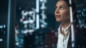 A woman in business attire stands by a window, contemplating her next job move. The window reflects city lights and blurred, glowing shapes, giving the scene a modern, introspective atmosphere.