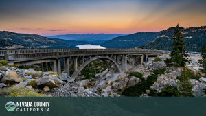 A scenic arched concrete bridge spans rocky terrain with a lake and forested mountains in the background at sunset, showcasing the natural beauty of Nevada County. The image includes a "Nevada County California" logo in the bottom left corner.