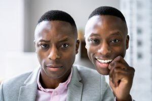 Two young men with short haircuts are posing closely together. The man on the left, channeling leadership, is wearing a light gray suit jacket and a pink shirt, looking serious. The man on the right, also dressed similarly, is smiling brightly while holding his hand near his face.