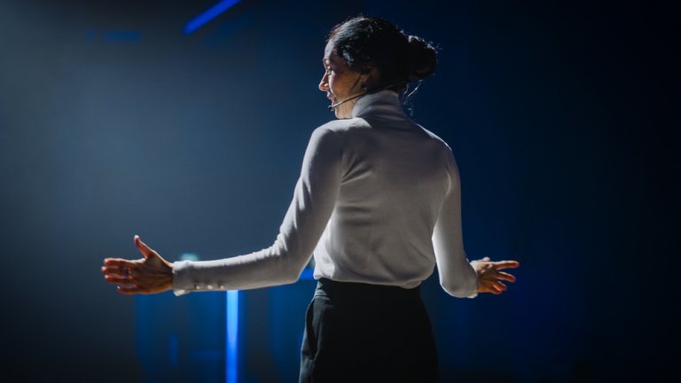 A woman presenting on a dim stage with lights focused on her